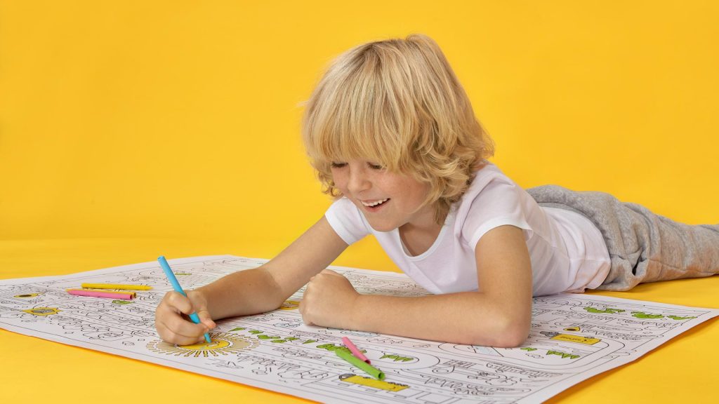 Zdjęcie przedstawia chłopca, który koloruje kredkami po dużej macie do kolorowania