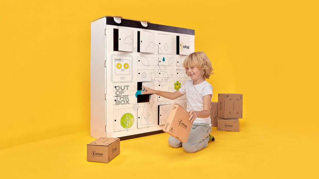Zdjęcie przedstawia chłopca, który trzyma w ręce paczkę przy otwartej skrytce w tekturowym Urządzeniu Paczkomat.