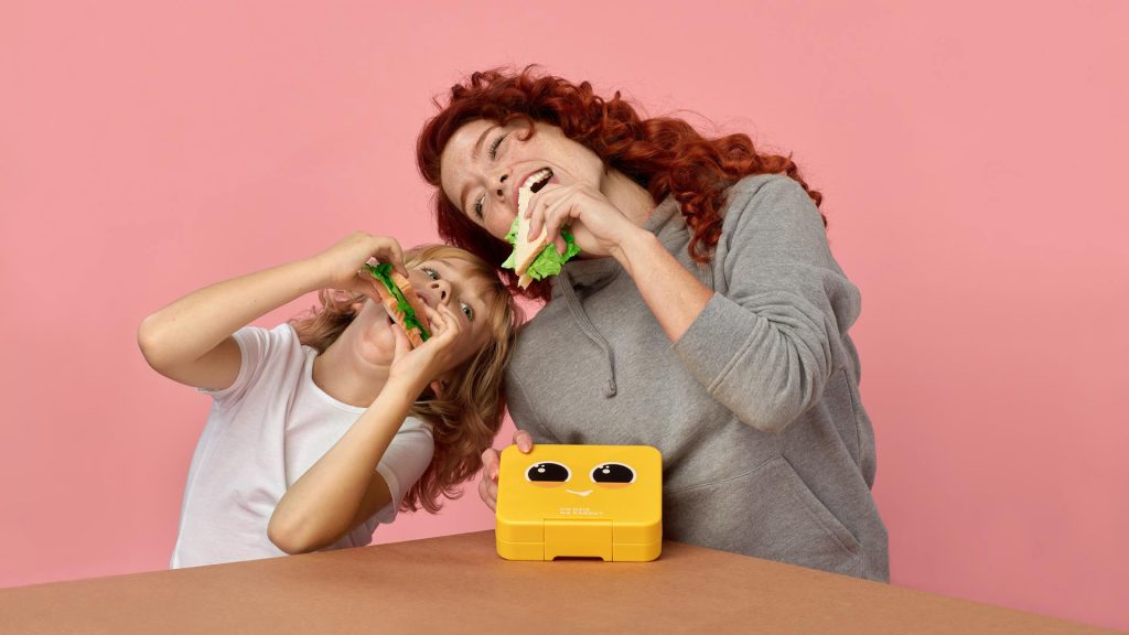 Zdjęcie przedstawia mamę z synem, którzy wygłupiając się jedzą kanapki z żółtego lunch boxa, który ma narysowane czarne oczy i biały nosek, przez co wygląda jak zabawna, animowana buźka.
