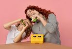Zdjęcie przedstawia mamę z synem, którzy wygłupiając się jedzą kanapki z żółtego lunch boxa, który ma narysowane czarne oczy i biały nosek, przez co wygląda jak zabawna, animowana buźka.