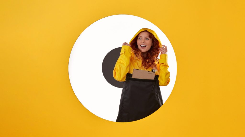 Zdjęcie przedstawia przeciwdeszczową kurtkę z kapturem, kaptur oraz góra kurtki wraz z rękawami jest żółta, a dalsza część kurtki, sięgająca do połowy uda jest czarna. W miejscu łączenia dwóch kolorów, na wysokości klatki piersiowej znajduje się duża kieszeń.