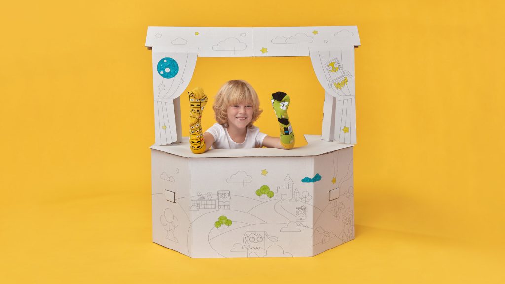 Zdjęcie przedstawia chłopca bawiącego się pacynkami w tekturowym teatrzyku.