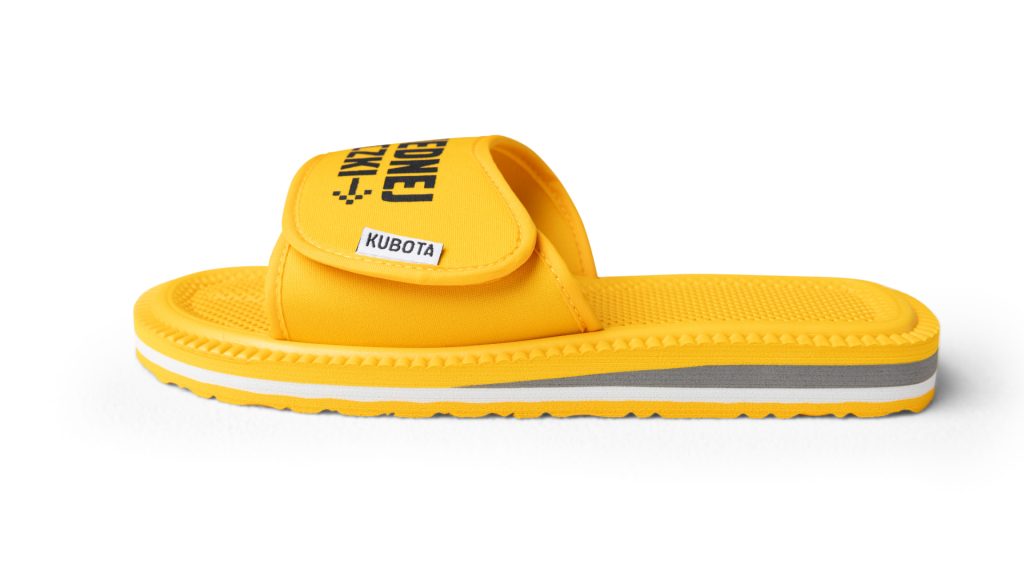 Zdjęcie przedstawia parę żółtych klapek marki Kubota na jasnozielonym tle. Na lewej klapce znajduje się napis „DWA LACZKI”, a na prawej „Z JEDNEJ PACZKI”. Klapki mają teksturę na podeszwie oraz szeroki pasek z zapięciem na rzep, na którym po bokach widnieje małe logo „KUBOTA”.