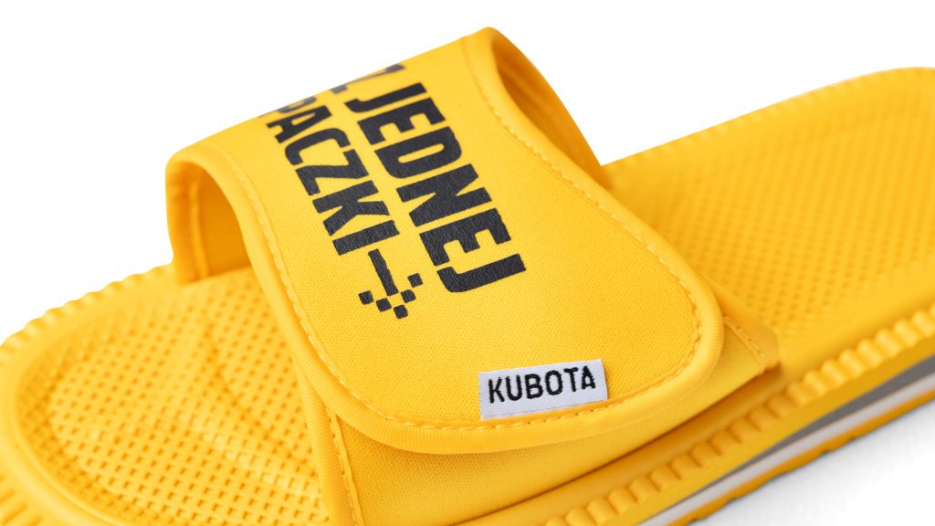 Zdjęcie przedstawia parę żółtych klapek marki Kubota na jasnozielonym tle. Na lewej klapce znajduje się napis „DWA LACZKI”, a na prawej „Z JEDNEJ PACZKI”. Klapki mają teksturę na podeszwie oraz szeroki pasek z zapięciem na rzep, na którym po bokach widnieje małe logo „KUBOTA”.