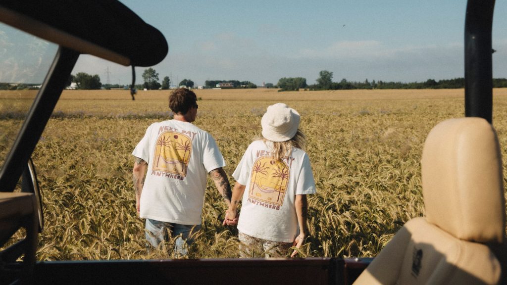 Para trzymająca się za ręce, mają na sobie białe t-shirty z nadrukiem maszyny Paczkomat, księżyco-słońcem na hamaku, który leży pod palmami.. T-shirt ma krótki rękaw.