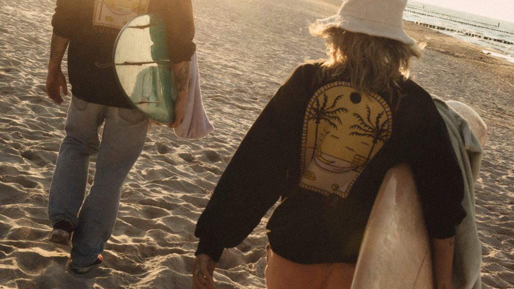 Surferzy idący plażą z deskami surfingowymi, ubrani w czarne bluzy.