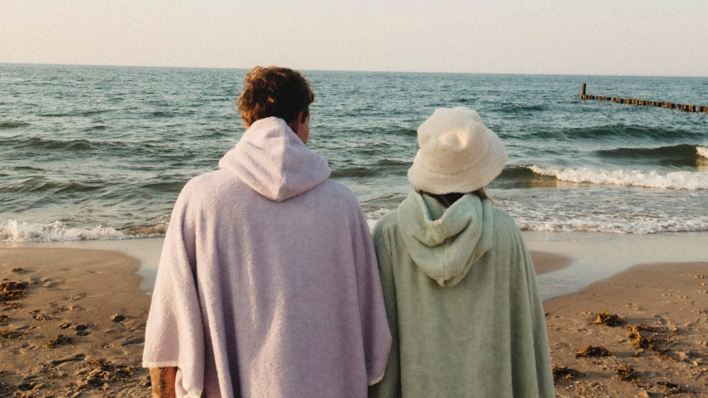 Dwie osoby w płaszczach na plaży, patrzą na morze.