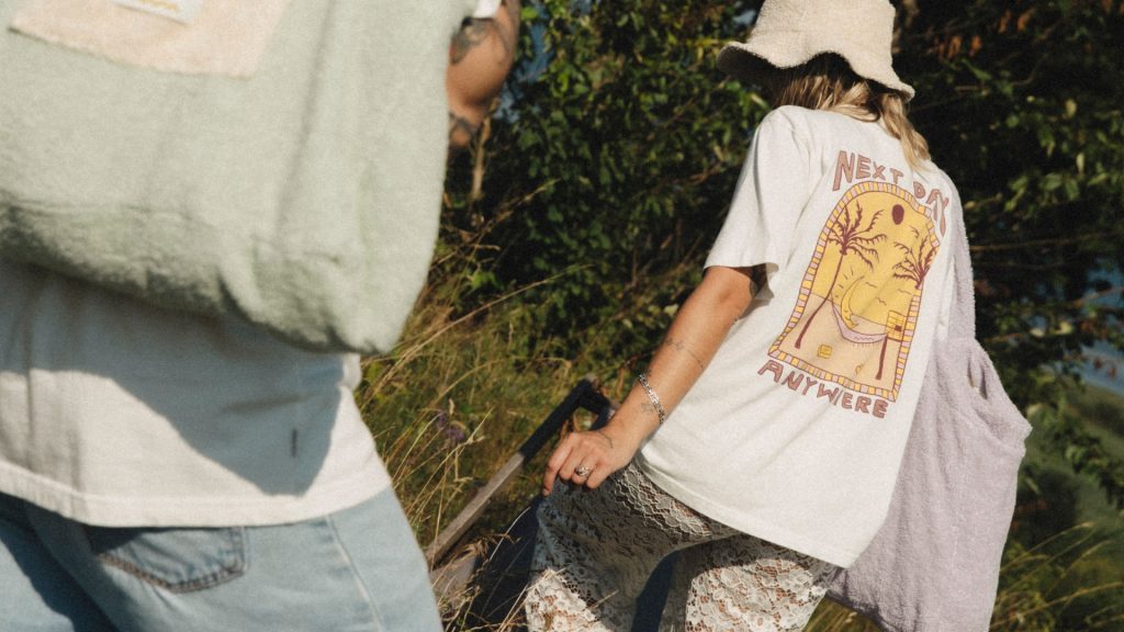 Dwie osoby w letnich ubraniach idą przez zarośla, jedna ma koszulkę z nadrukiem.