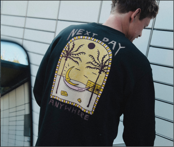 Meżczyzna wkłada paczkę do maszyny Paczkomat. Ma na sobie czarną bluzę z dużym żółto-brązowo-białym nadrukiem na plecach, przedstawiającym rajską plaże z półksięzycem na hamaku, na tle Paczkomatu. Nadruk jest opatrzony napisem "next day anywhere"