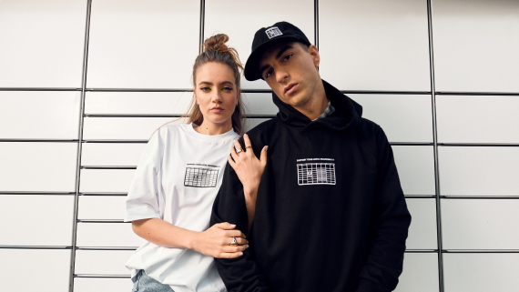 Para stojaca na tle maszyny Paczkomat. Mają na sobie ubrania z kolekcji Support Your local paczkomat. On ma czarną bluzę z napisem Support your local paczkomat oraz z maszyna Paczkomat. Ona ma biały t-shirt.