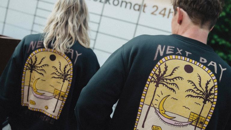 Para w czarnych bluzach z nadrukiem "Next Day Anywhere" stoją przed maszyną Paczkomat